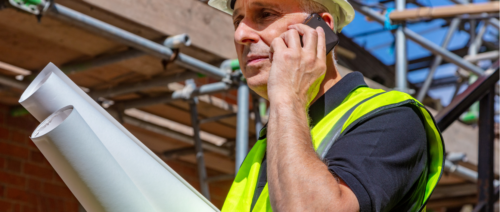 Site Manager on phone holding blueprints in a construction site
